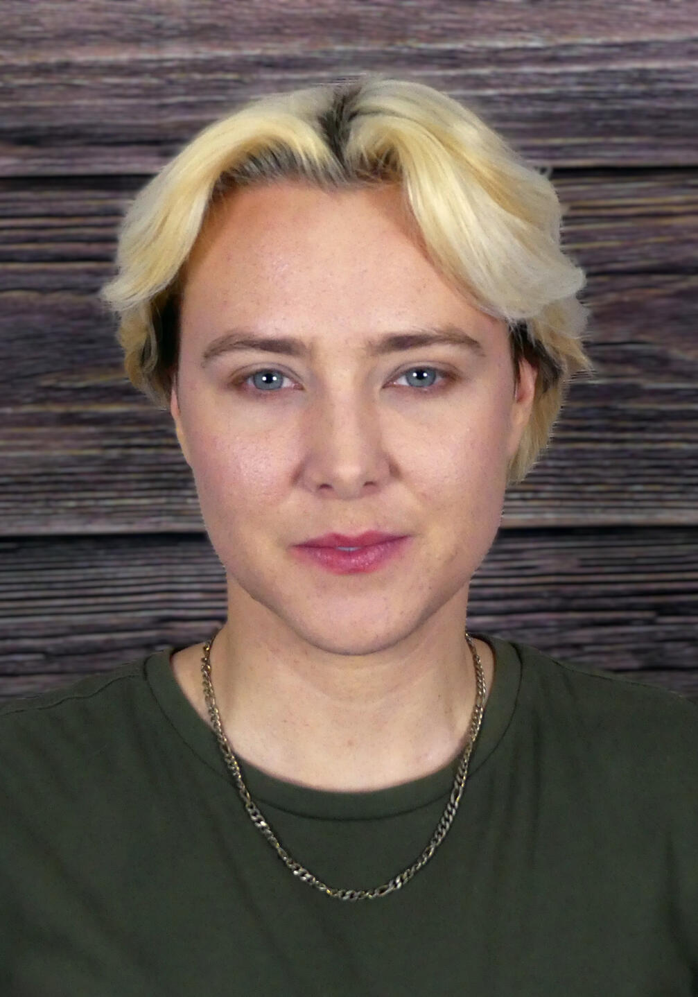 Hayden's Headshot: A white person with bleach-blonde hair and blue eyes, wearing a muted green shirt and a gold necklace. Blurred wood background.