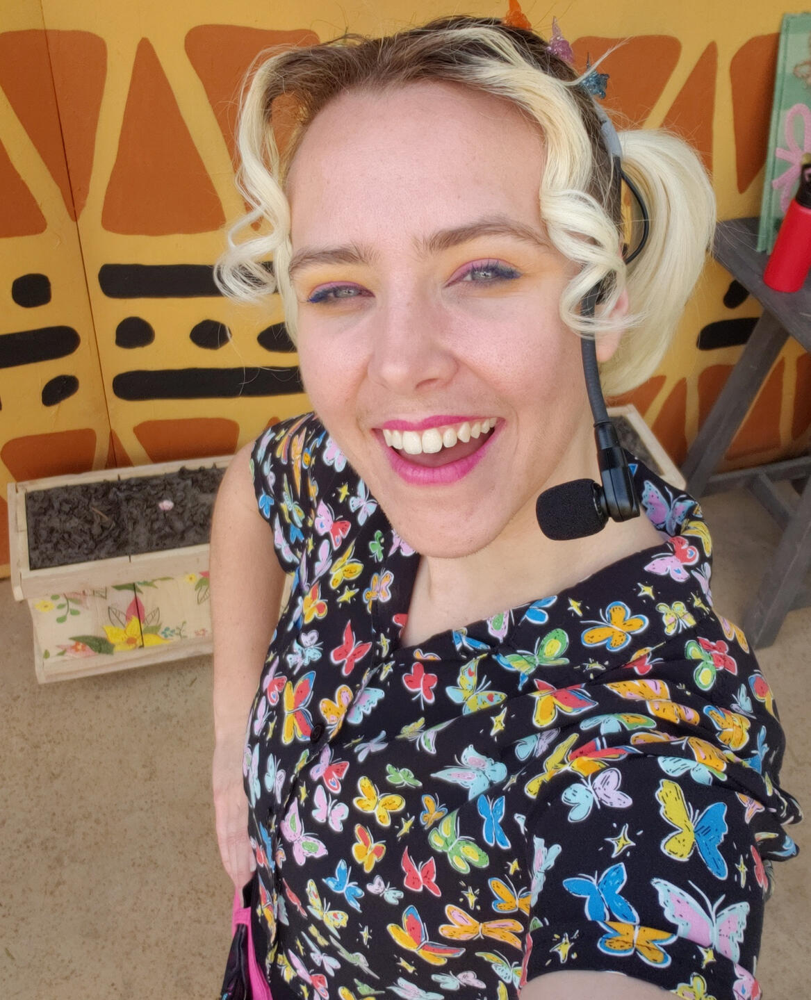 A selfie of Hayden at the Safari Park with curly blonde hair, a microphone headset, and colorful butterflies on their shirt.