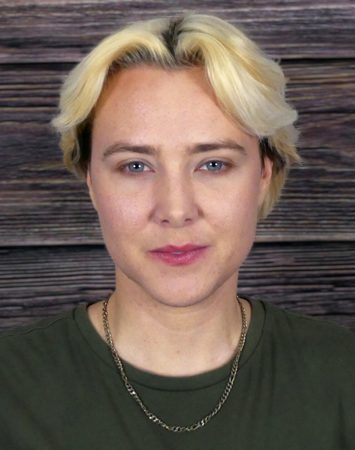 Hayden's Headshot: A white person with bleach-blonde hair and blue eyes, wearing a muted green shirt and a gold necklace. Blurred wood background.